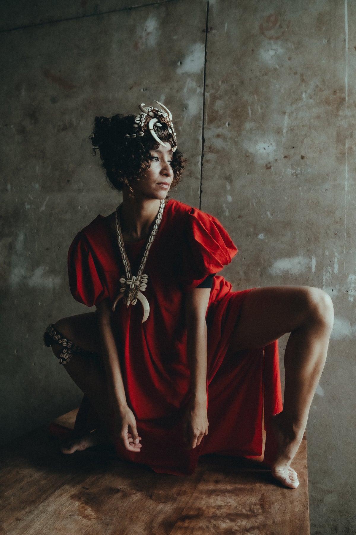 Model seated in the red Sexy Talar with shell jewellery against a textured wall.